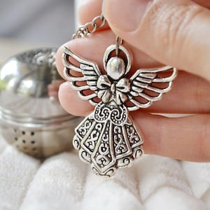 Puede incluir: Infusor de té de ángel de plata con un diseño detallado y una cadena. El infusor está siendo sostenido por una mano.