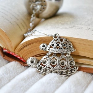 May include: A silver tea infuser with a chain and a decorative turtle charm. The charm is detailed with geometric patterns and small black accents. The infuser is resting on an open book.