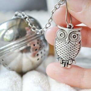May include: Silver tea infuser with a chain and an owl-shaped charm. The infuser is a sphere with holes and the owl charm has detailed feathers and large eyes.