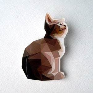Peut inclure: Figurine de chat géométrique dans les tons marron, beige et blanc. Le chat est assis, regardant vers le haut. Le design est composé de formes triangulaires, lui donnant un aspect moderne et artistique. La figurine est placée sur un fond blanc.