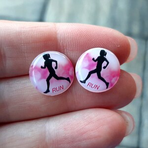Puede incluir: Un par de pendientes redondos blancos con un fondo de acuarela rosa. Cada pendiente presenta una silueta negra de un corredor y la palabra "RUN" impresa en negro.
