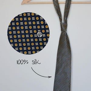 Pode incluir: Gravata de seda azul marinho com um padr&atilde;o repetido de pequenos quadrados e c&iacute;rculos amarelos e brancos. A gravata est&aacute; pendurada em um cabide de madeira. Um close-up mostra o detalhe do tecido e o texto "100% seda".