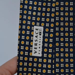 Pode incluir: Uma gravata azul marinho com um padr&atilde;o repetido de pequenas formas florais amarelas e brancas. A gravata tem uma etiqueta que diz "VERSACE CLASSIC V2". Um acess&oacute;rio cl&aacute;ssico.