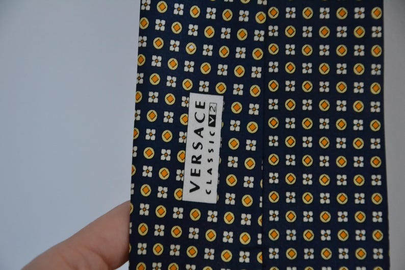 Pode incluir: Uma gravata azul marinho com um padr&atilde;o repetido de pequenas formas florais amarelas e brancas. A gravata tem uma etiqueta que diz "VERSACE CLASSIC V2". Um acess&oacute;rio cl&aacute;ssico.