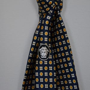 Pode incluir: Gravata de seda azul marinho com um padr&atilde;o repetido de pequenas formas florais amarelas e brancas. A gravata apresenta um medalh&atilde;o branco com um logotipo de cabe&ccedil;a de Medusa. Um acess&oacute;rio cl&aacute;ssico para trajes formais ou de neg&oacute;cios.