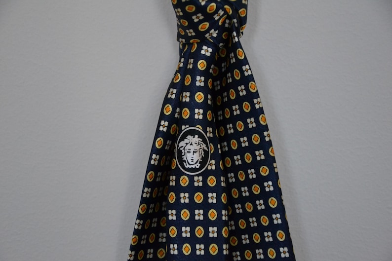 Pode incluir: Gravata de seda azul marinho com um padr&atilde;o repetido de pequenas formas florais amarelas e brancas. A gravata apresenta um medalh&atilde;o branco com um logotipo de cabe&ccedil;a de Medusa. Um acess&oacute;rio cl&aacute;ssico para trajes formais ou de neg&oacute;cios.