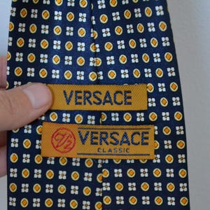 Pode incluir: Gravata Versace azul marinho com um padr&atilde;o repetido de pequenas formas florais brancas e laranja. A gravata tem duas etiquetas douradas com o nome da marca "VERSACE" e "VERSACE CLASSIC".