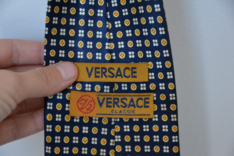 Pode incluir: Gravata Versace azul marinho com um padr&atilde;o repetido de pequenas formas florais brancas e laranja. A gravata tem duas etiquetas douradas com o nome da marca "VERSACE" e "VERSACE CLASSIC".
