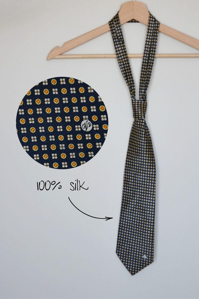 Pode incluir: Gravata de seda azul marinho com um padr&atilde;o repetido de pequenos quadrados e c&iacute;rculos amarelos e brancos. A gravata est&aacute; pendurada em um cabide de madeira. Um close-up mostra o detalhe do tecido e o texto "100% seda".