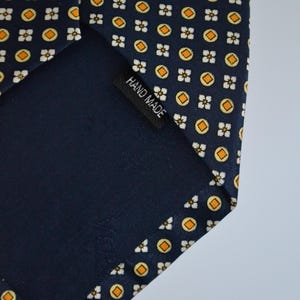 Pode incluir: Gravata azul marinho com um padr&atilde;o repetido de c&iacute;rculos laranja e formas florais brancas. A gravata tem um verso azul escuro e uma etiqueta que diz "HAND MADE".