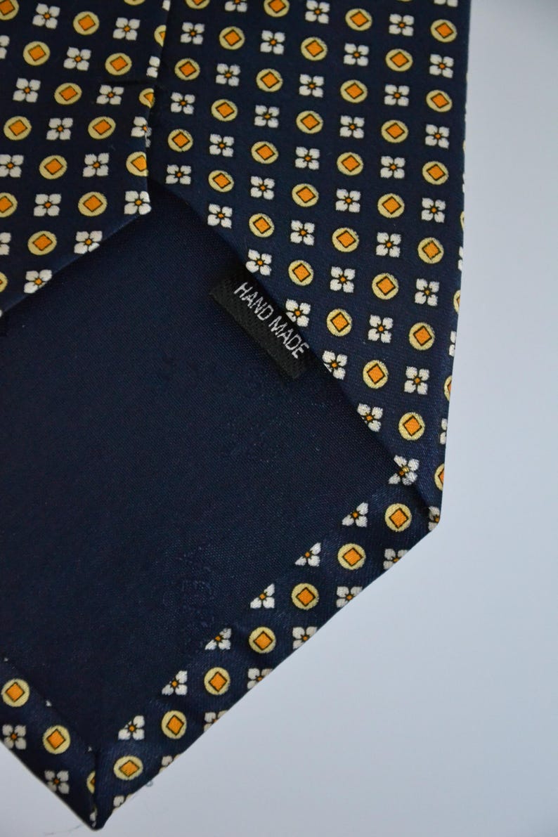 Pode incluir: Gravata azul marinho com um padr&atilde;o repetido de c&iacute;rculos laranja e formas florais brancas. A gravata tem um verso azul escuro e uma etiqueta que diz "HAND MADE".