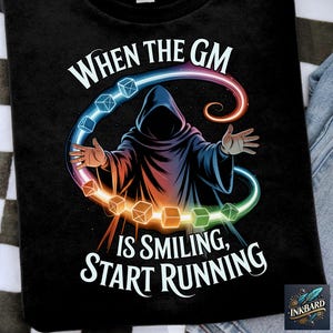 Op de afbeelding: Zwart t-shirt met een fantasieontwerp. Het toont een figuur met een capuchon met uitgestrekte handen, omringd door gloeiende dobbelstenen en een kleurrijke boog. Tekst luidt: "When the GM is Smiling, Start Running".