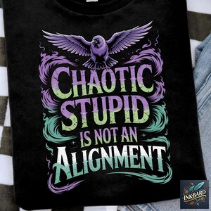 May include: Black t-shirt featuring the phrase "CHAOTIC STUPID IS NOT AN ALIGNMENT" in purple, green, and white. A purple bird with spread wings is above the text. The shirt is on a checkered surface.
