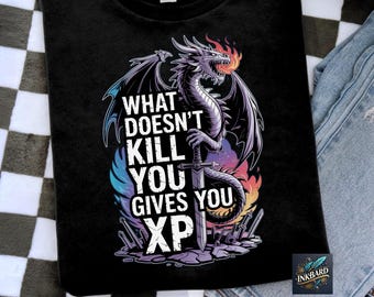 Op de afbeelding: Zwart T-shirt met een afbeelding van een paarse draak die een zwaard vasthoudt. De tekst "What doesn't kill you gives you XP" staat op het T-shirt gedrukt.
