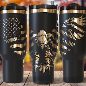 May include: Three black travel mugs with gold accents and handles. The mugs feature patriotic designs: an American flag, an eagle, and eagle wings. The mugs are on a wooden surface.
