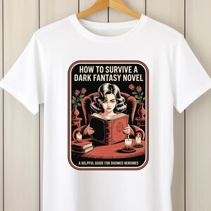 A white t-shirt with a graphic design depicting a woman reading a book, surrounded by various objects such as candles, vases, and a potted plant.