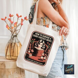 A woman is standing next to a wooden stool, holding a white tote bag with a graphic design on it. The bag features a woman reading a book and the text "How to Survive a Dark Fantasy Novel" printed on it.