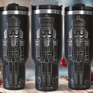 May include: Three black travel mugs with a silver nutcracker design. Each mug has a handle and a lid. The nutcracker is depicted in detail, with a hat, uniform, and gift boxes at the bottom. The mugs are suitable for hot or cold beverages.