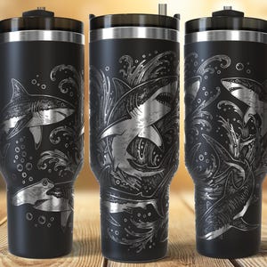 Puede incluir: Taza de viaje de acero inoxidable negro con un diseño de tiburón. La taza tiene un asa y una tapa. El diseño del tiburón está grabado en plata, con olas y burbujas. La taza está diseñada para mantener las bebidas calientes o frías.
