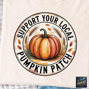 May include: Cream-colored t-shirt featuring a circular design with a pumpkin illustration. The design includes the text "SUPPORT YOUR LOCAL PUMPKIN PATCH" in a black font, with wheat stalks and fall leaves. The shirt is made of a soft material.