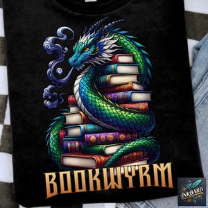 Op de afbeelding: Een zwart T-shirt met een groene en blauwe draak die om een stapel boeken is gewikkeld. De draak heeft een gouden kroon en de tekst "BOOKWORM" is in gouden letters onder de draak gedrukt.