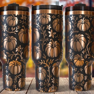 May include: Three black travel mugs with a gold pumpkin and floral design. Each mug has a black handle and lid with a gold rim. The background is a blurred autumn scene.