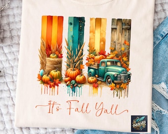 May include: Cream-colored t-shirt featuring a watercolor design with fall elements. The design includes pumpkins, autumn leaves, and a teal pickup truck. Text reads "It's Fall Y'all" in an orange script. The design is set against colorful vertical stripes.