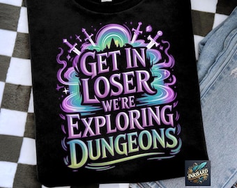 Op de afbeelding: Zwart t-shirt met een kleurrijke afbeelding met de tekst "Get in Loser We're Exploring Dungeons". Het ontwerp toont zwaarden, een regenboog en een berglandsilhouet. Het shirt wordt getoond op een geruit oppervlak.