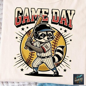 Può includere: T-shirt color crema con un giocatore di baseball procione che tiene una mazza, su uno sfondo di baseball e raggi di stelle. Il testo "GAME DAY" è sopra il procione e "Trash Panda" sulla maglia.