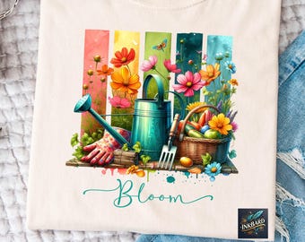 Puede incluir: Una camiseta blanca con un gráfico colorido de flores, una regadera, herramientas de jardinería y una cesta de verduras. El gráfico está rodeado de un efecto acuarela. El texto "Bloom" está escrito en turquesa debajo del gráfico.