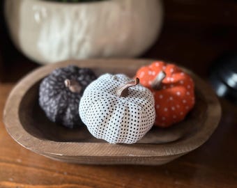 May include: Three fabric pumpkins in a wooden bowl. The pumpkins are in black and white polka dot, white and black polka dot, and orange with white polka dots. The pumpkins have brown stems and are a fall decoration.