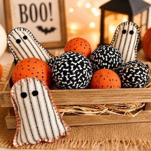 May include: A wooden crate filled with fabric decorations including three ghost-shaped figures made of cream and black striped fabric with black felt eyes, and several round fabric balls featuring orange with black dots or black with white bone patterns.