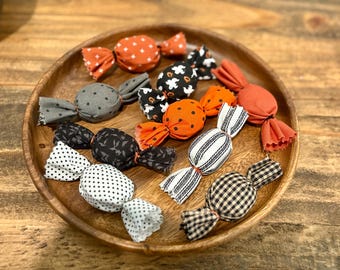 May include: A wooden bowl holds several fabric candies in various patterns and colors. The candies are wrapped with fabric in orange, black, white, and gray, featuring polka dots, stripes, and Halloween-themed designs. The bowl sits on a wooden surface.