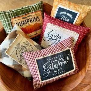 May include: A collection of five small, rectangular fabric pillows in a wooden bowl. The pillows feature various patterns including green plaid, red gingham, and mustard yellow floral, each with a central patch displaying text like Farm-Fresh Pumpkins, Bless, Give Thanks, Gather, and Grateful.