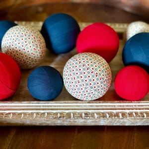 May include: A collection of decorative spheres in a wooden tray. The spheres are in various sizes and colors, including red, navy blue, and patterned with a red and blue star design. The tray has a distressed, white-washed finish.