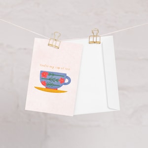 May include: A pink card with the text "You're my cup of tea" and a blue teacup with red flowers and green leaves on a yellow saucer. The card is attached to a white envelope with gold binder clips.