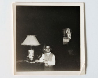 May include: Black and white photo of a young boy standing in front of a table with a lamp. The boy is wearing a light-colored shirt and a dark vest. The lamp has a white shade and a decorative base.