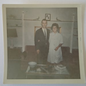 May include: Vintage photograph of a couple posing indoors. The man wears a dark suit and tie, while the woman is in a light-colored dress. A coffee table with magazines and a decorative bowl is in front of a red sofa.