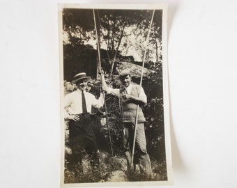 May include: Black and white photograph of two men standing in a wooded area. Both men are wearing hats and holding long sticks. The man on the left is wearing a white shirt and tie, while the man on the right is wearing a sweater.