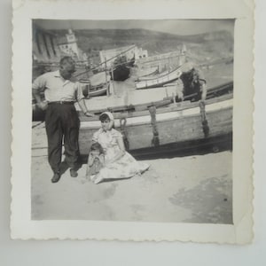 Può includere: Una foto in bianco e nero di una famiglia in piedi vicino a una barca su una spiaggia. La famiglia è composta da un uomo, una donna e un bambino piccolo. L'uomo indossa una camicia bianca e pantaloni scuri. La donna indossa un vestito e un foulard. Il bambino indossa un vestito. La barca è di legno e ha una corda legata intorno.