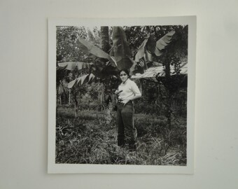 May include: A black and white photograph of a woman standing in front of a banana tree. She is wearing a white shirt and jeans. The photo was taken outdoors.