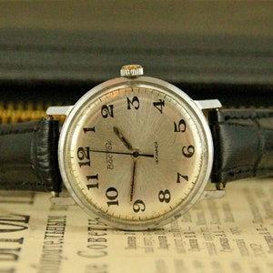 May include: A silver wristwatch with a black leather band. The watch face has black Roman numeral hour markers and a silver second hand. The watch is on a newspaper.