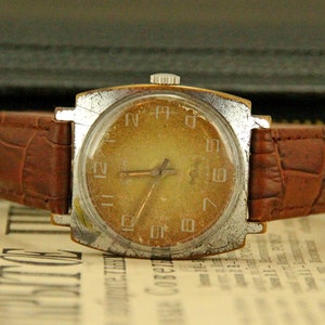 May include: A vintage square-shaped wristwatch with a brown leather band. The watch face is cream-colored with black Roman numeral hour markers and black hands.