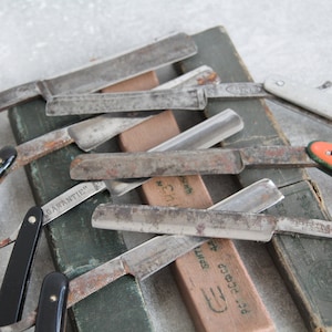 Puede incluir: Una colección de navajas de afeitar rectas vintage con varios mangos, incluyendo negro, blanco y naranja. Las navajas están dispuestas sobre una caja de madera con una etiqueta verde que dice "P.G. ECCLES & SMITH" y "GUARANTIE" en alemán.