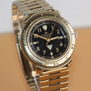 May include: A gold-tone wristwatch with a black face and a gold-tone metal band. The watch face has white markings and the text "CARDI VOSTOK" and "GP" printed on it.