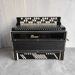 Puede incluir: Un acordeón negro con ribetes plateados y teclas blancas. El acordeón tiene la palabra "Bemo" escrita en letras doradas en la parte delantera.