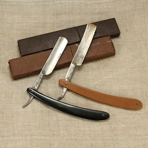 Puede incluir: Dos navajas de afeitar rectas antiguas con mangos y estuches marrones y negros. Las navajas son de metal y tienen una hoja afilada.