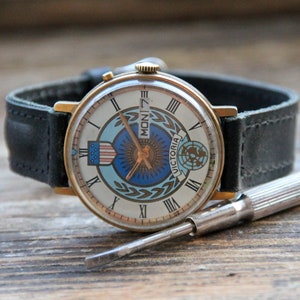 May include: A gold-tone wristwatch with a white face and black leather band. The face features a blue and white globe design with the word "VICTORIA" and the day of the week "MON 7".