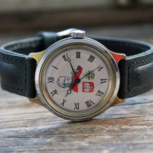 May include: A vintage wristwatch with a white face and black hands. The face features Roman numerals and a small image of a man in a red and white uniform. The watch has a black leather band.