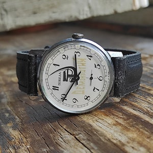 May include: A black leather wristwatch with a white face and black hands. The watch face has the word "Победа" (Victory) and a stylized image of a rocket ship. The numbers on the watch face are in black.